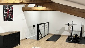 Une vaste pièce mansardée avec un plafond en bois, une porte en métal noir, un espace dédié à la musculation et un tapis aux motifs géométriques.