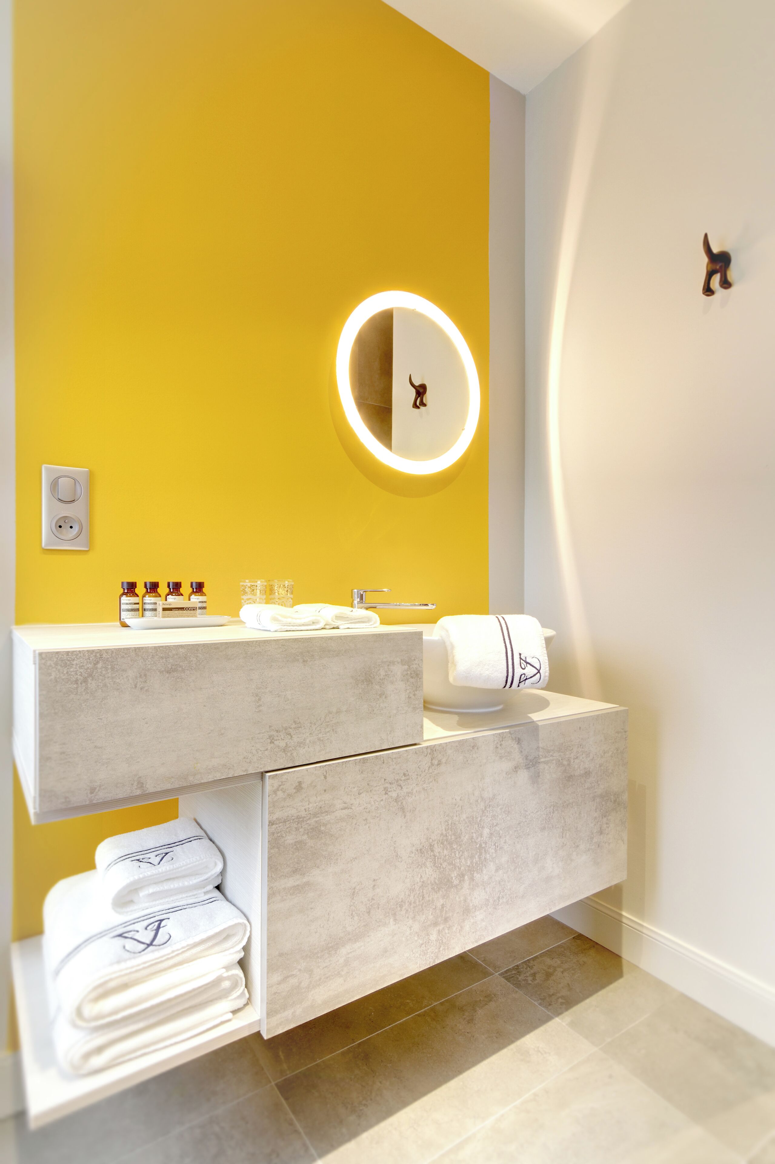 Un baño moderno con una pared de acento amarilla, un espejo grande con luz circular, tocador de mármol y toallas dobladas con cuidado.