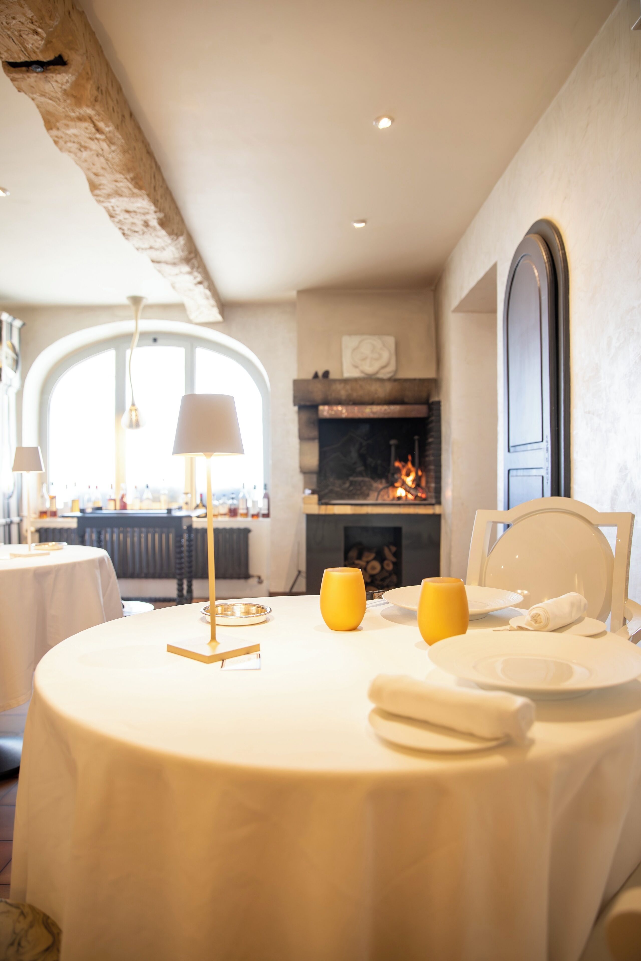 Un comedor con una mesa redonda preparada para dos, una chimenea al fondo y vistas a través de un amplio ventanal en arco.