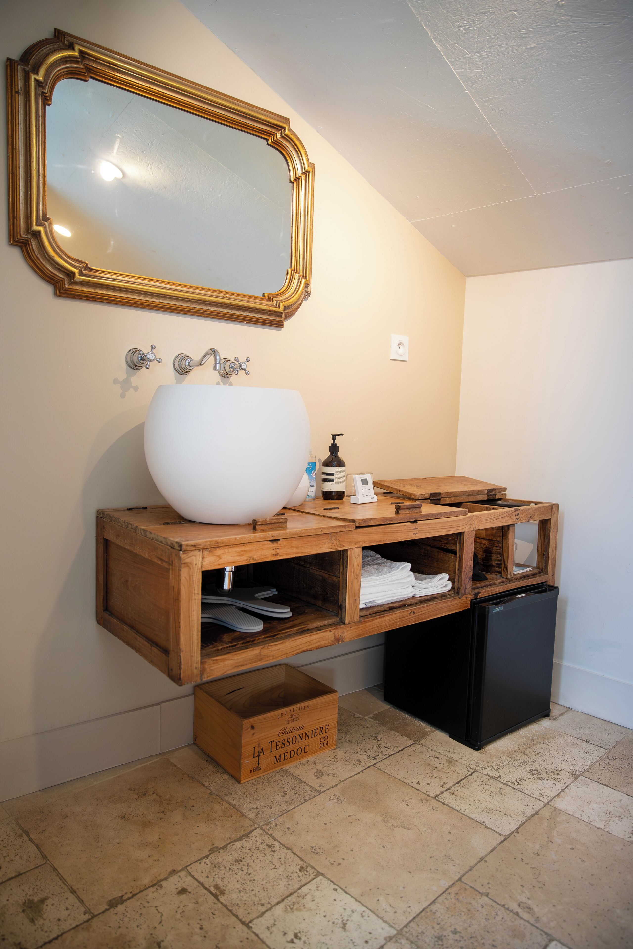 Un baño con un lavabo grande blanco, tocador de madera, un espejo con marco dorado y un refrigerador negro.