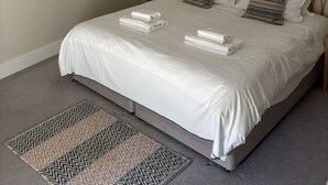 A neatly made bed with white linens and two folded towels, a simple white pendant light, and a framed landscape painting on the wall.