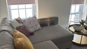 A living room with a grey sectional sofa, patterned cushions, a round coffee table, and a framed picture on the wall.