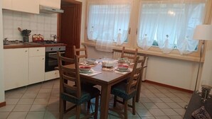 A dining area with a wooden table set for four, surrounded by chairs. There is a kitchen area with a stove and cabinets to the left.