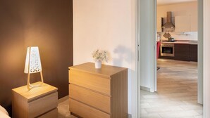 A bedroom with a wooden dresser, a bedside lamp, and a view into the kitchen through an open door. - Suite 2 (Casamassima)