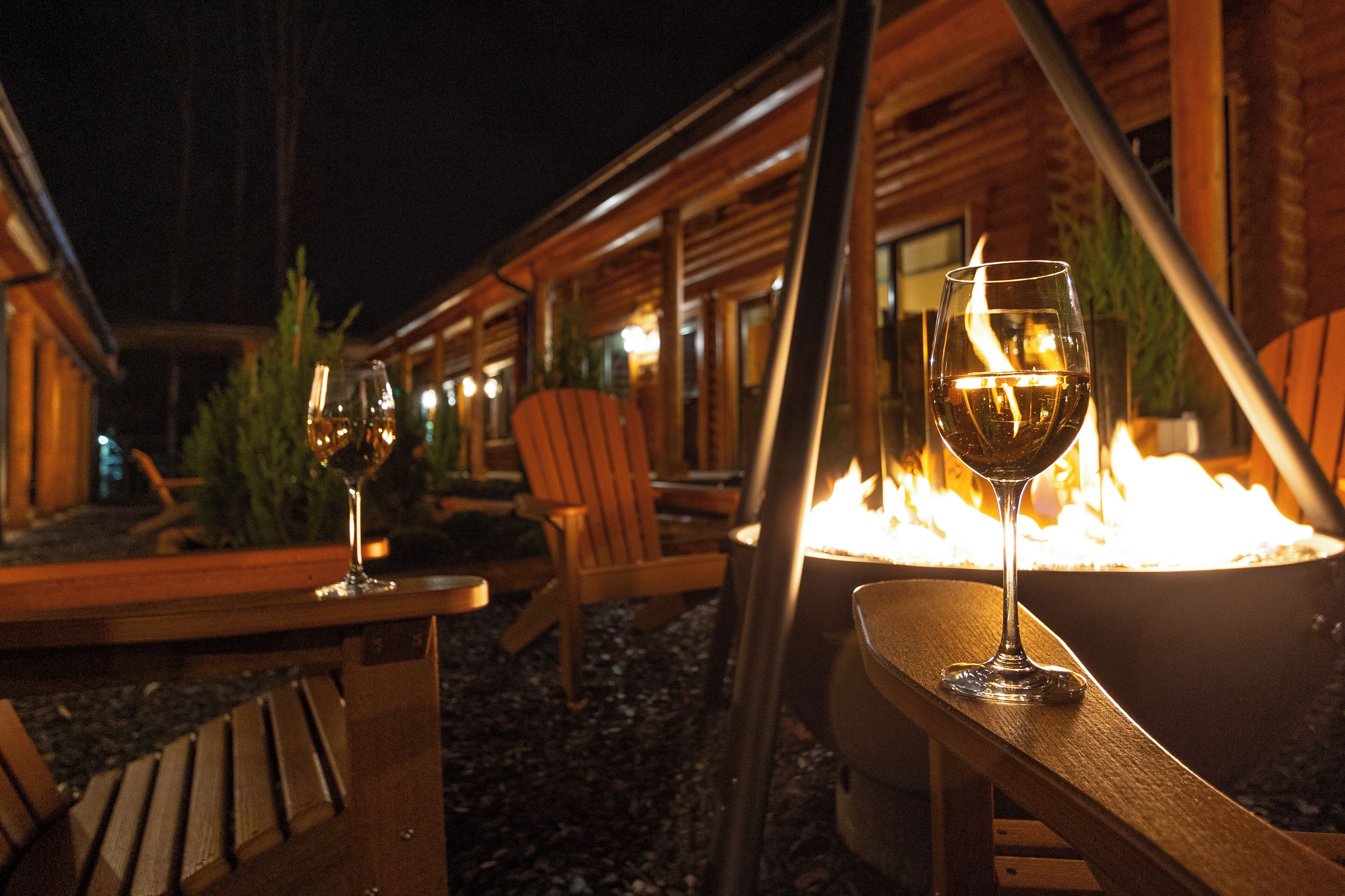 A wooden deck with two tables, each with a glass of wine and a chair. A large fire pit is visible in the background.