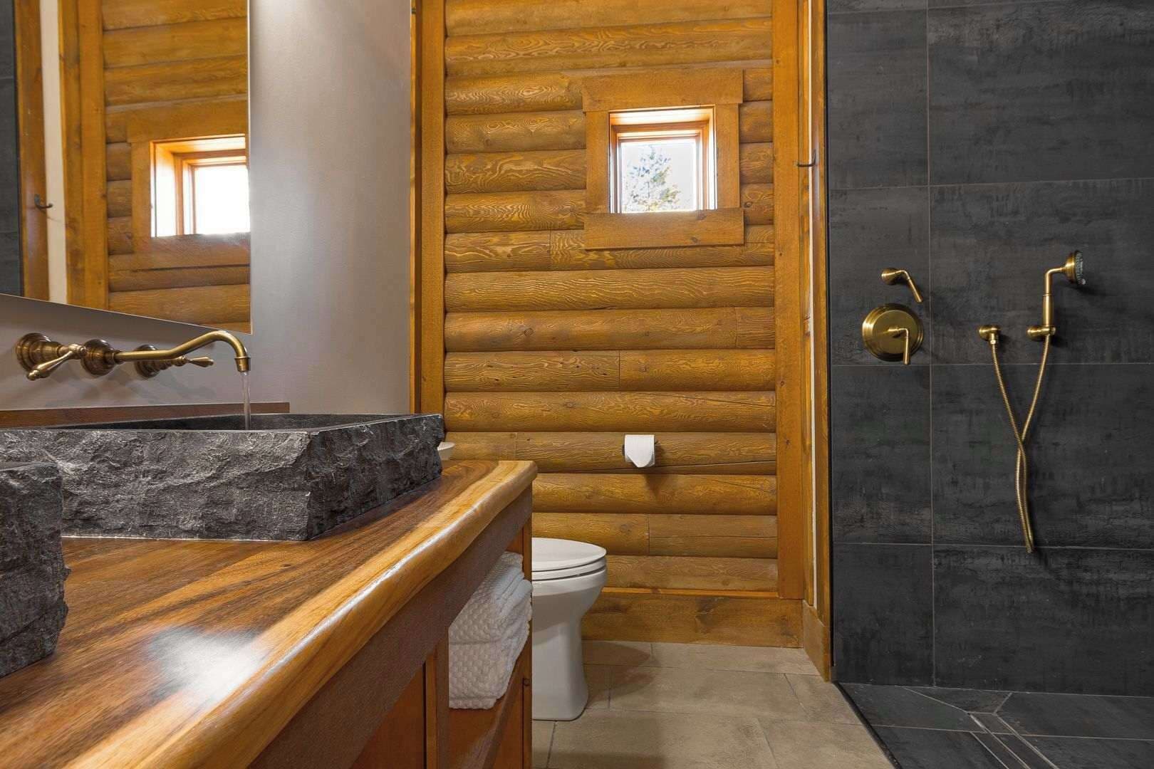 A bathroom with a wooden log wall, a stone sink, a toilet, and a shower with brass fixtures.
