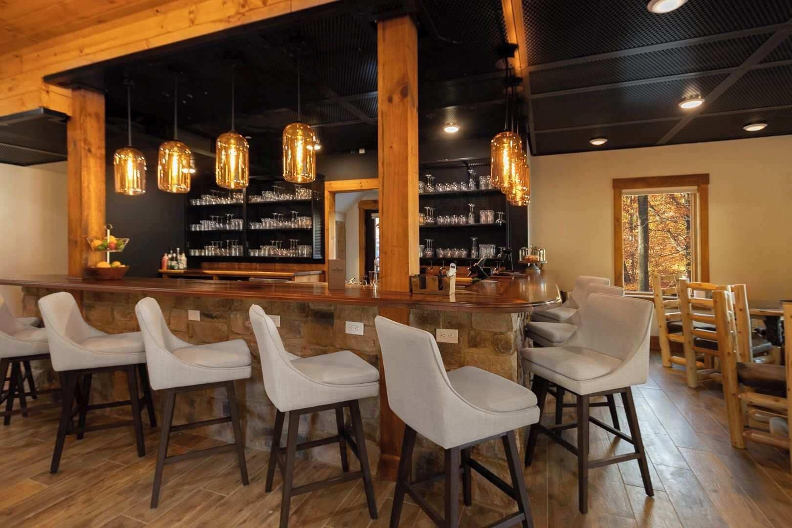 Zona de bar con taburetes de madera, lámparas colgantes y estantes con copas y botellas.