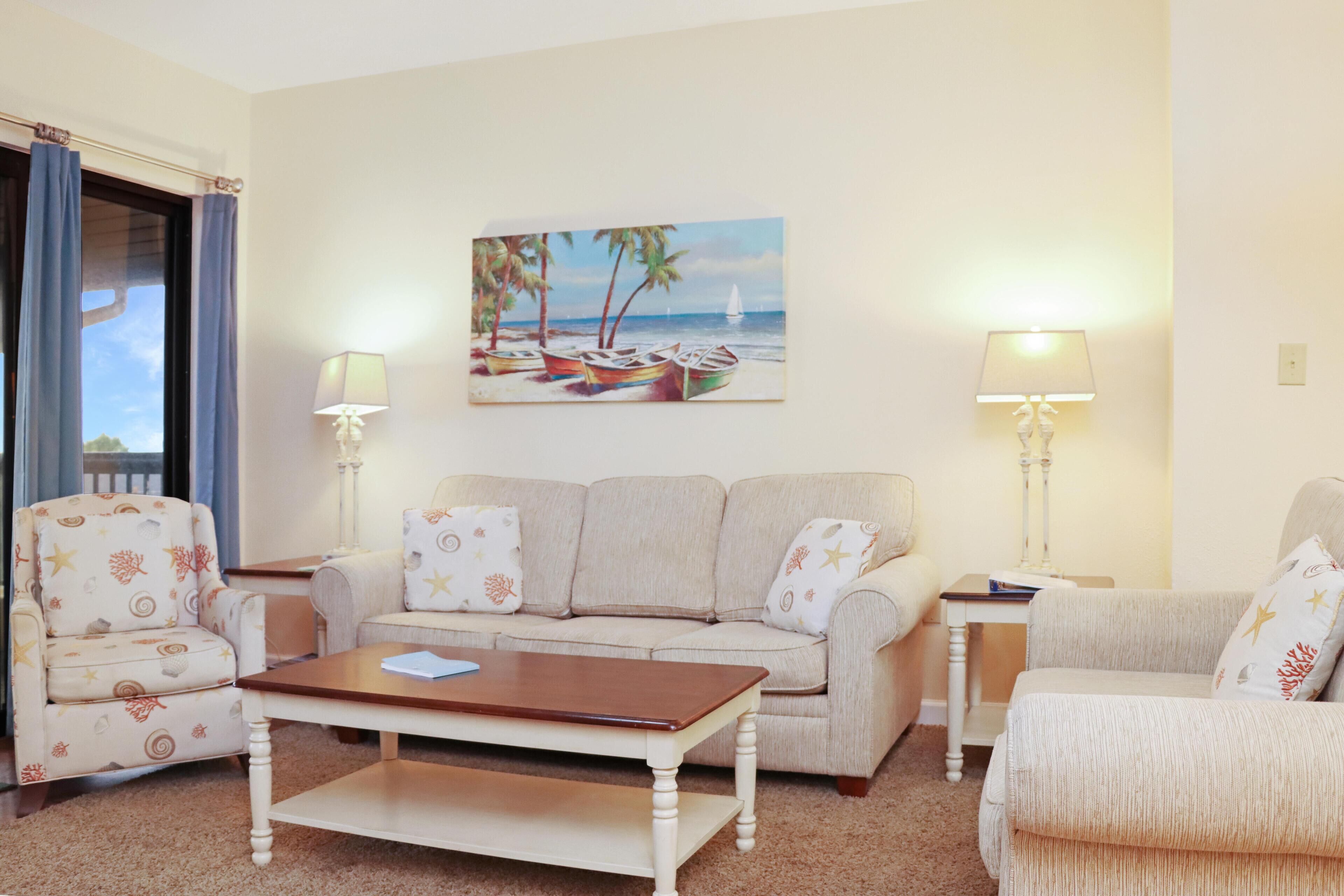 A living room with a beige sofa, two armchairs, a coffee table, and a painting of a beach scene. There are two lamps on side tables.