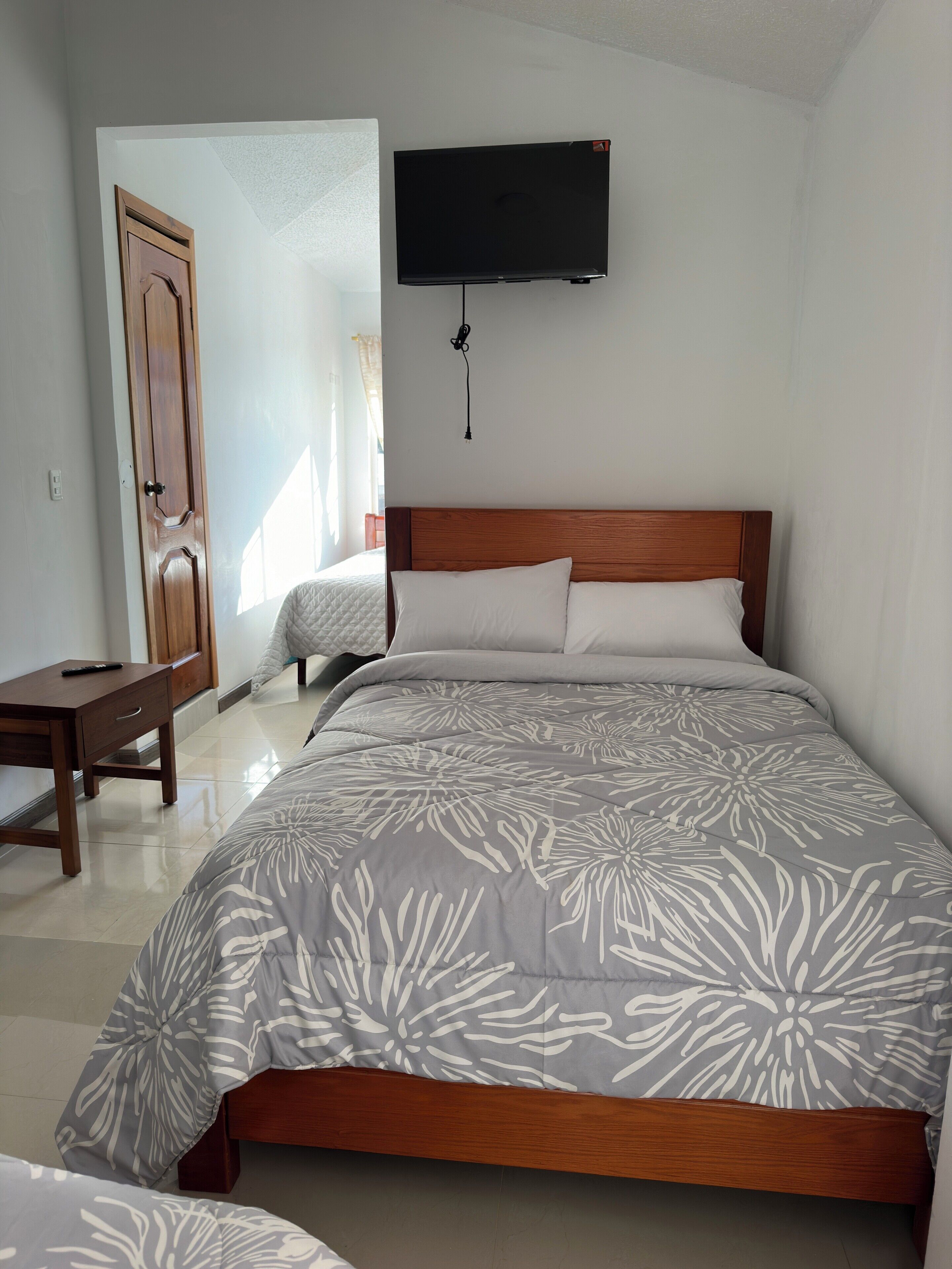 Een slaapkamer met een houten bed, een flatscreen televisie aan de muur, een houten nachtkastje en een deur die leidt naar een andere kamer.