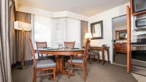 A hotel room with a dining table, chairs, a lamp, a TV, and a kitchenette.