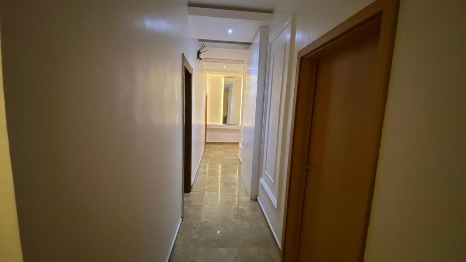 A narrow hallway with a wooden door on the right, a white wall on the left, and a light source at the end.