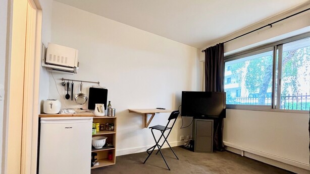 A compact room with a kitchenette, a foldable chair, a flat-screen TV, and a window with a view of trees and a fence. - 5 min to Parc des Princes • Studio 18 m²<br>Boulogne (Boulogne-Billancourt)