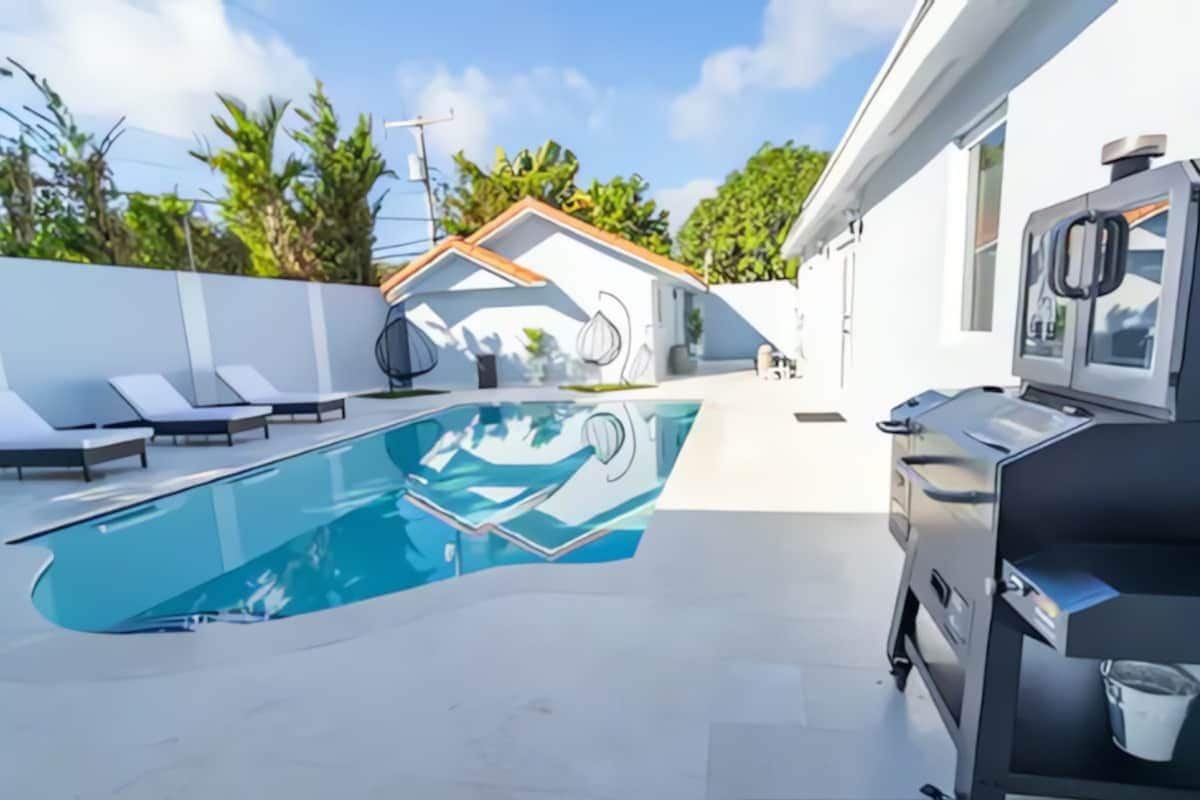 A swimming pool with lounge chairs, a barbecue grill, and a white building with a tiled roof.