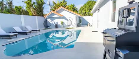 Une piscine avec des chaises longues, un barbecue et un bâtiment blanc doté d’un toit en tuiles.