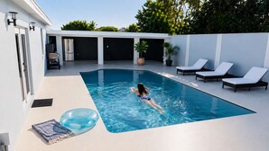 Une piscine où une personne nage, des chaises longues et un barbecue.