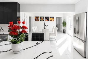 A modern kitchen with a white marble countertop, a stainless steel refrigerator, and a dining area with white chairs and a white table.