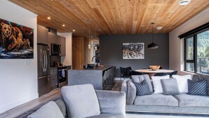 A modern living space with a wooden ceiling, grey sofas, a kitchen area, and artwork on the walls.