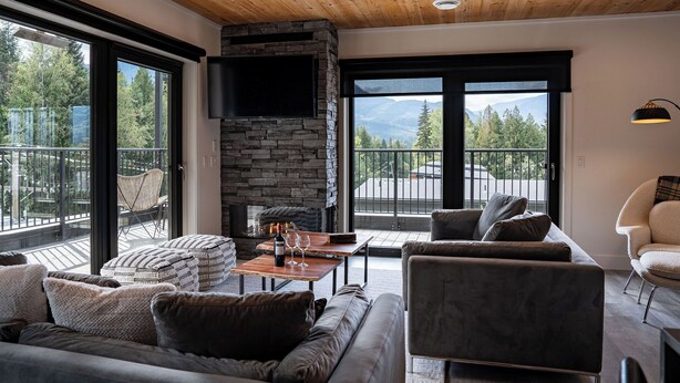 A modern living room with a stone fireplace, large windows, and a balcony with a view of the mountains.