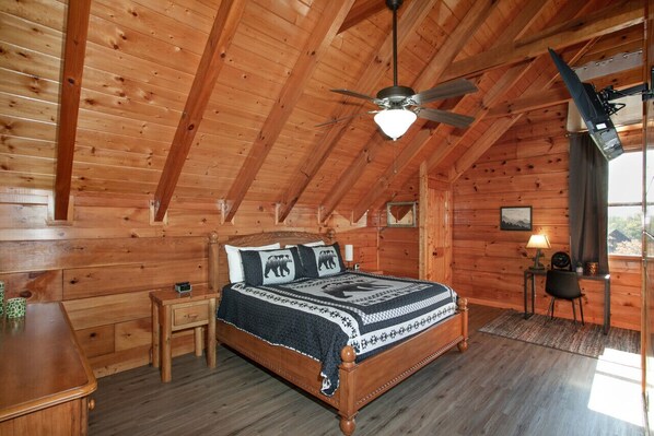 Une chambre dans une cabane en bois, avec un lit, un bureau, une chaise, un ventilateur de plafond et une fenêtre.