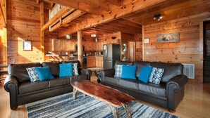 Ein Holzblockhaus-Interieur mit einem Wohnzimmer, das zwei Sofas mit gemusterten Kissen, einen Holztisch und einen im Hintergrund sichtbaren Küchenbereich umfasst.
