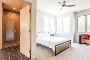 A bedroom with a bed, a desk, a chair, a ceiling fan, and a shower area.