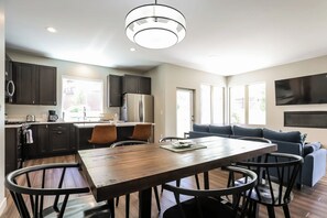 A modern kitchen with dark cabinets, a central island with chairs, a dining table, and a large window providing natural light.