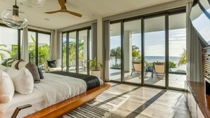 Un dormitorio moderno con una cama grande, un ventilador de techo y vista al océano a través de ventanales de piso a techo.