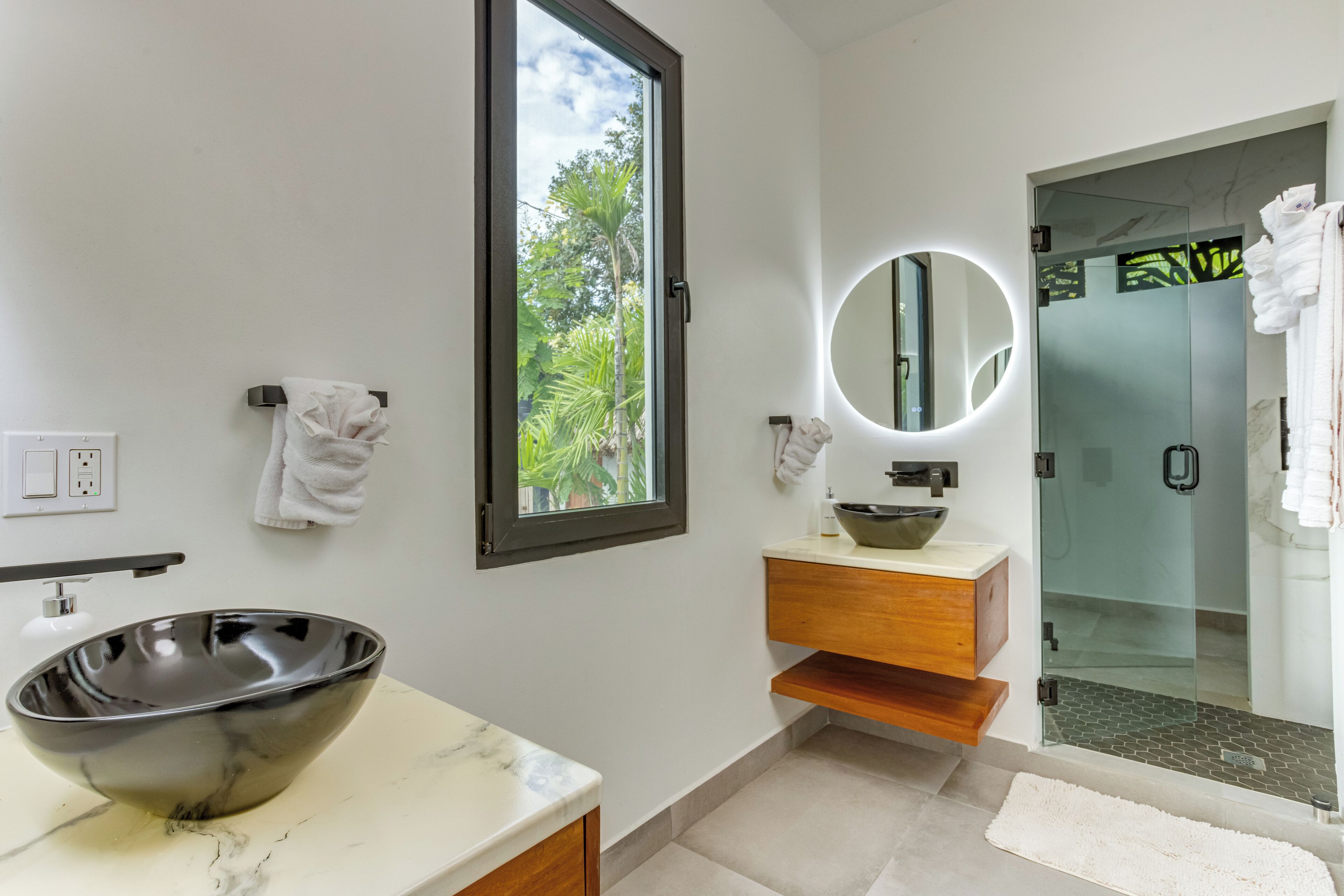 A modern bathroom with a large bowl sink, a wooden vanity, a glass shower enclosure, and a view of greenery outside.