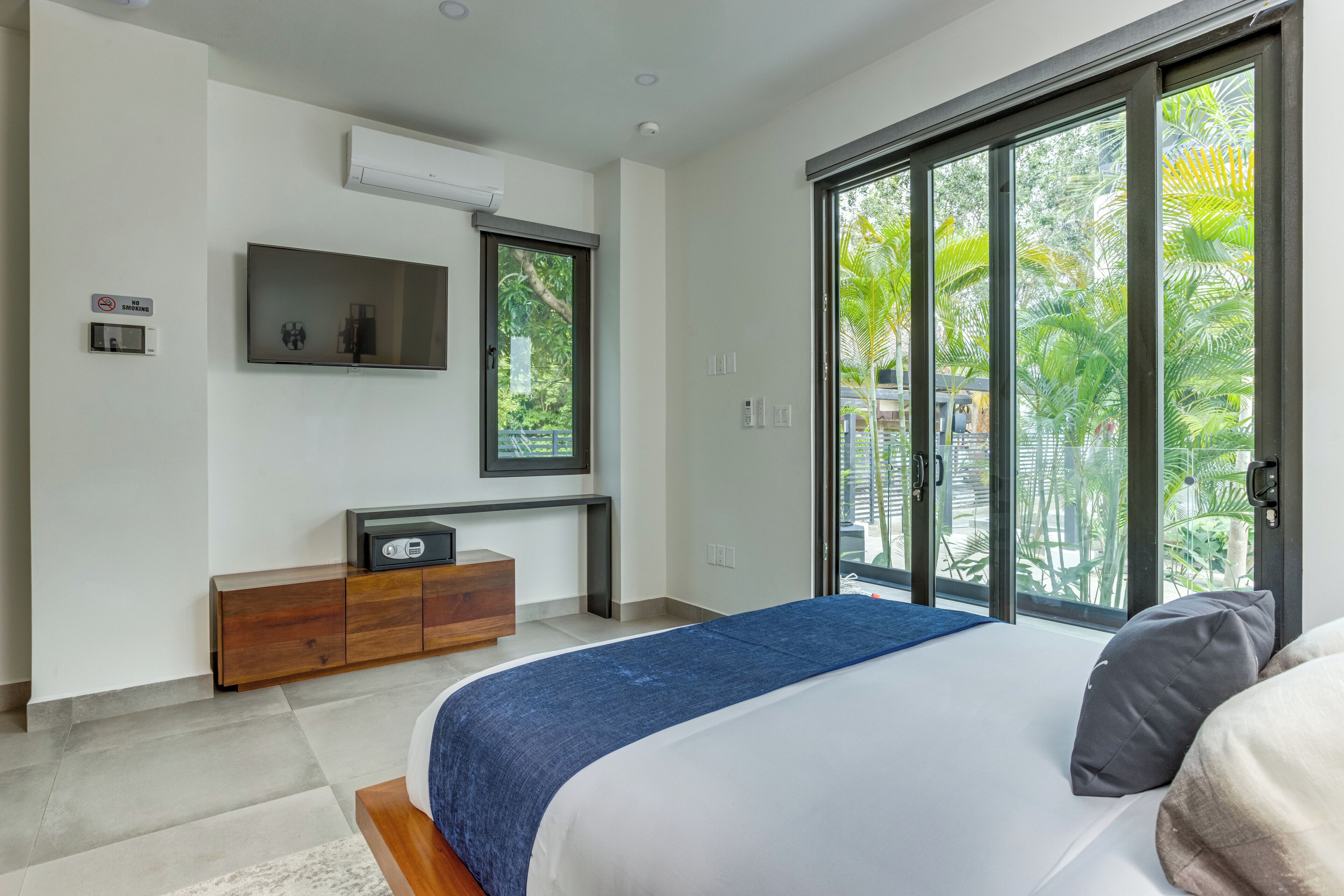 Un dormitorio moderno con una cama grande, un televisor en la pared y un ventanal corredero que da a un balcón con plantas.