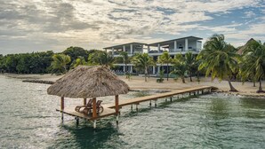 Una propiedad frente a la playa con una terraza de madera, una estructura de techo de paja y un edificio moderno con varios balcones.