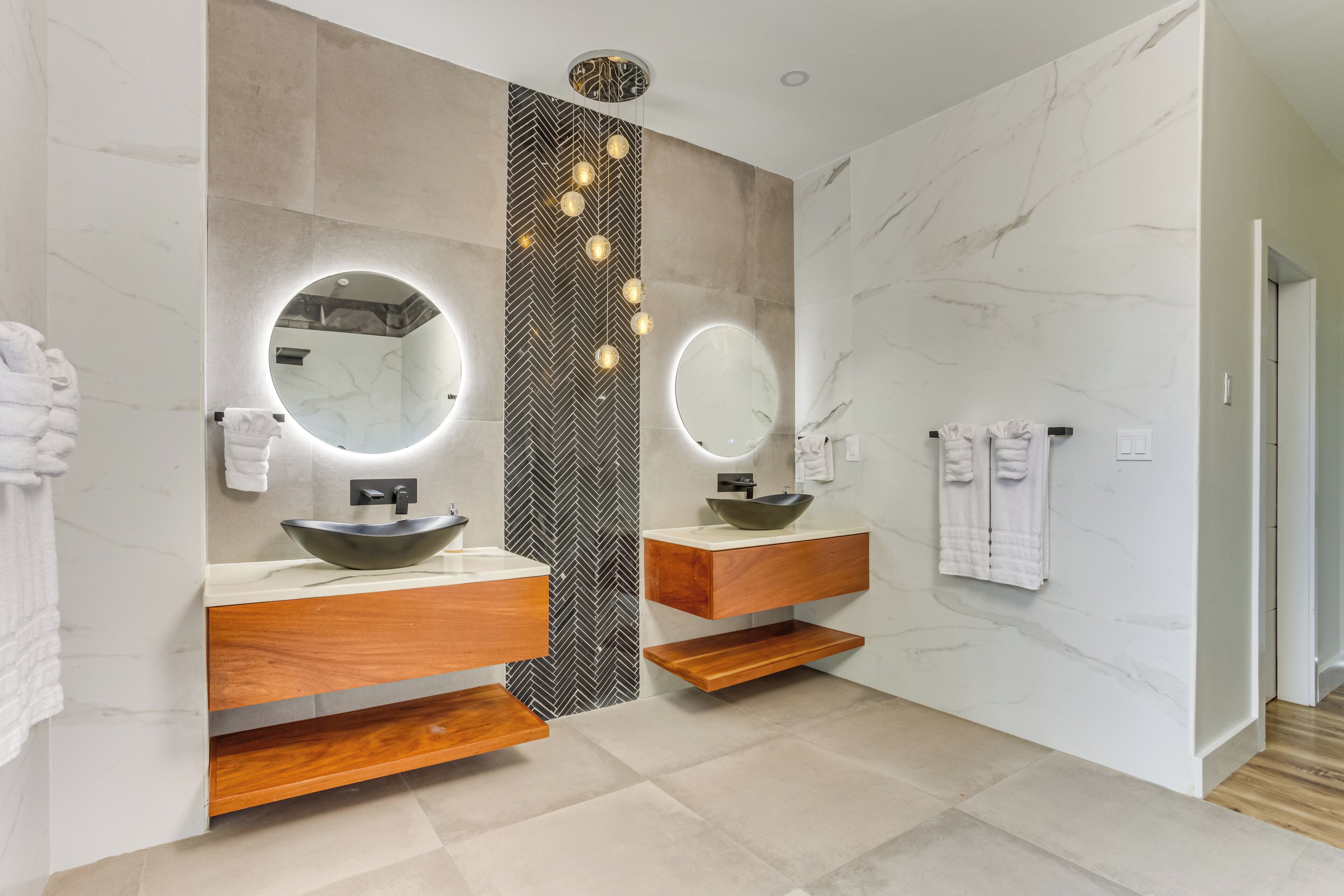 Un baño moderno con dos lavamanos, tocador de madera, paredes de mármol y una pared de acento con diseño de espiga.