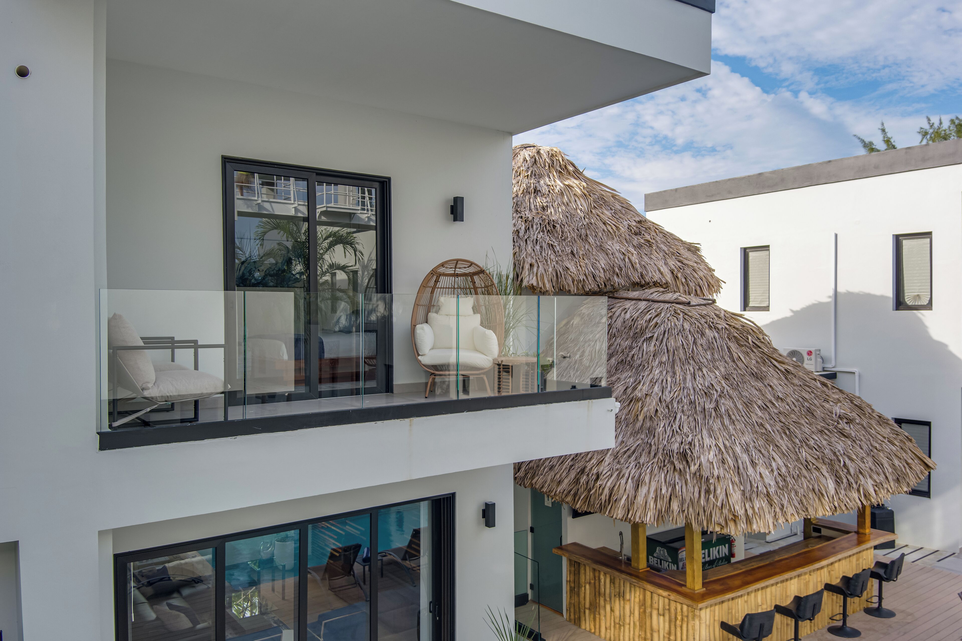 Un edificio moderno de dos pisos con un balcón de vidrio, un área de estar al aire libre con techo de paja y una barra de madera.