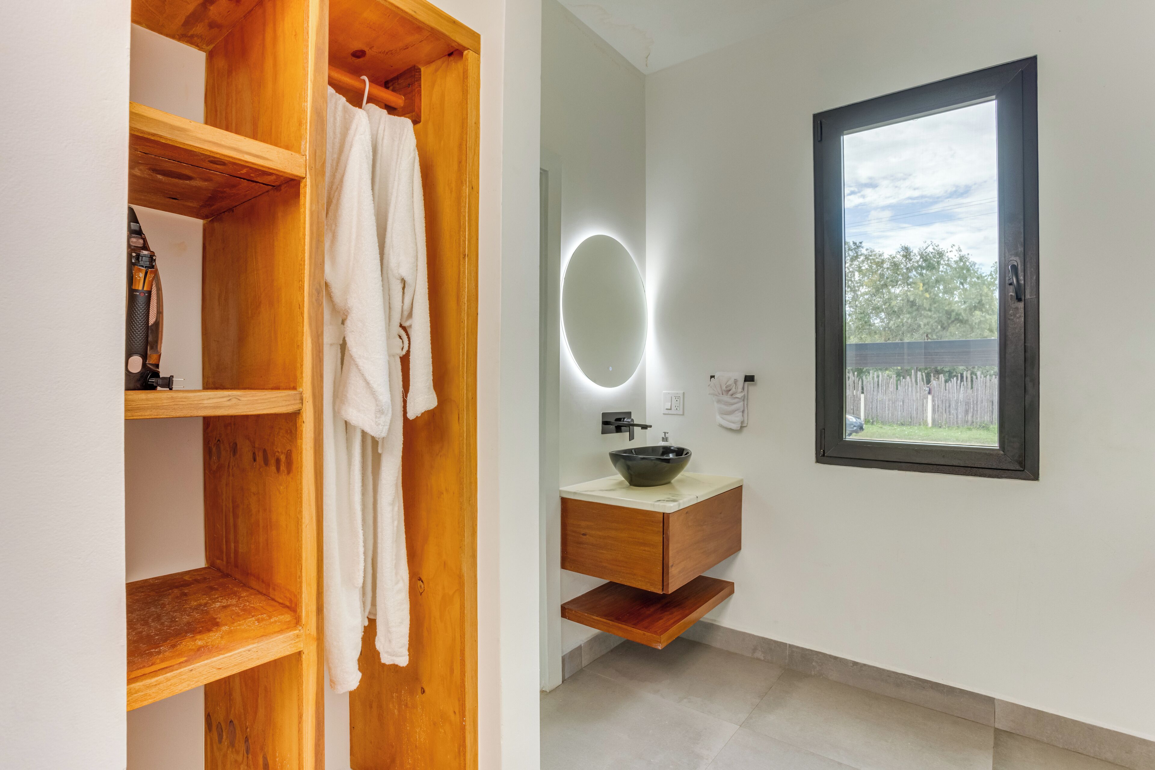 A bathroom with a wooden vanity, a round mirror with lights, a window, and a towel rack.