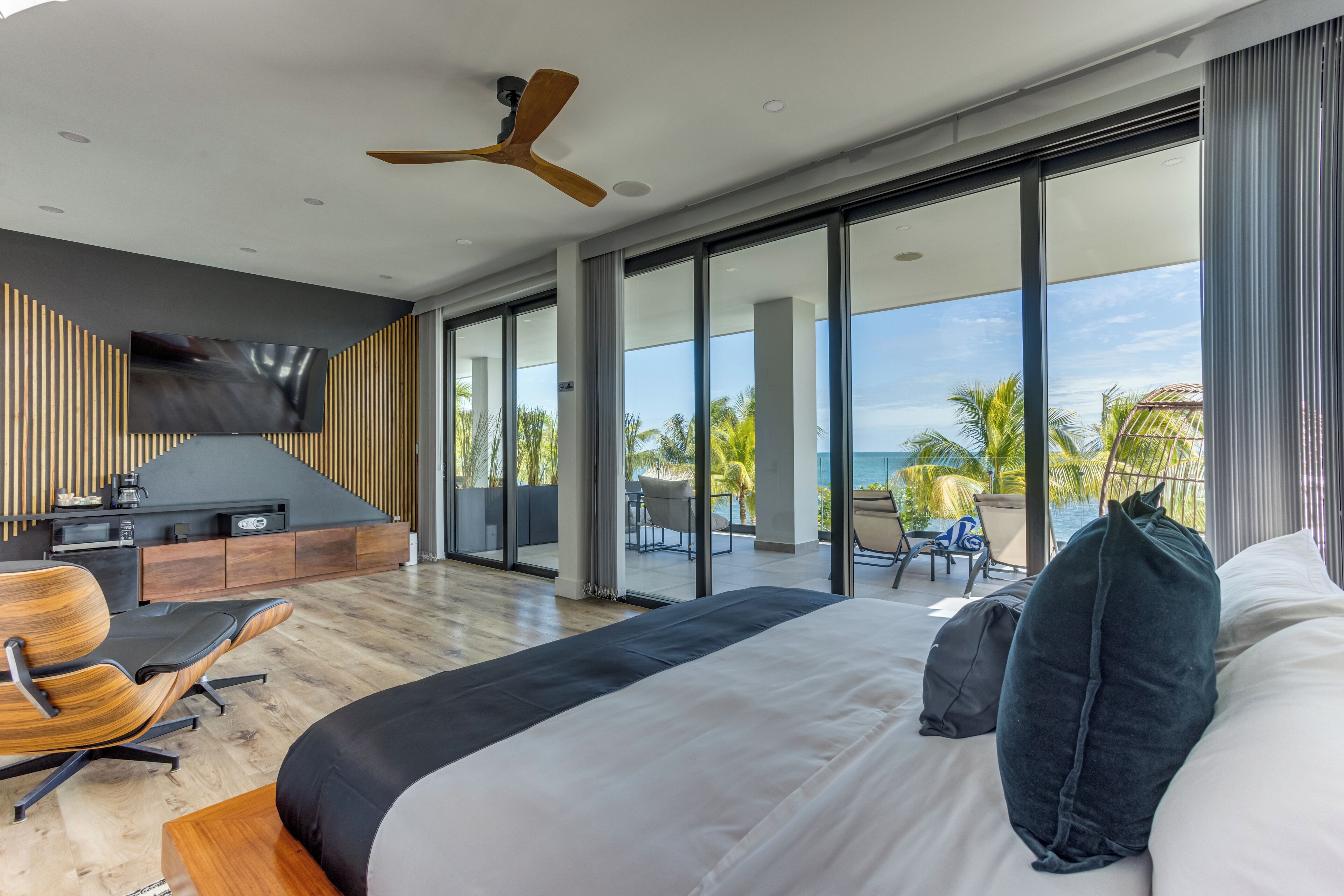 A modern hotel room with a large bed, a TV, a ceiling fan, and a balcony with a view of the ocean.