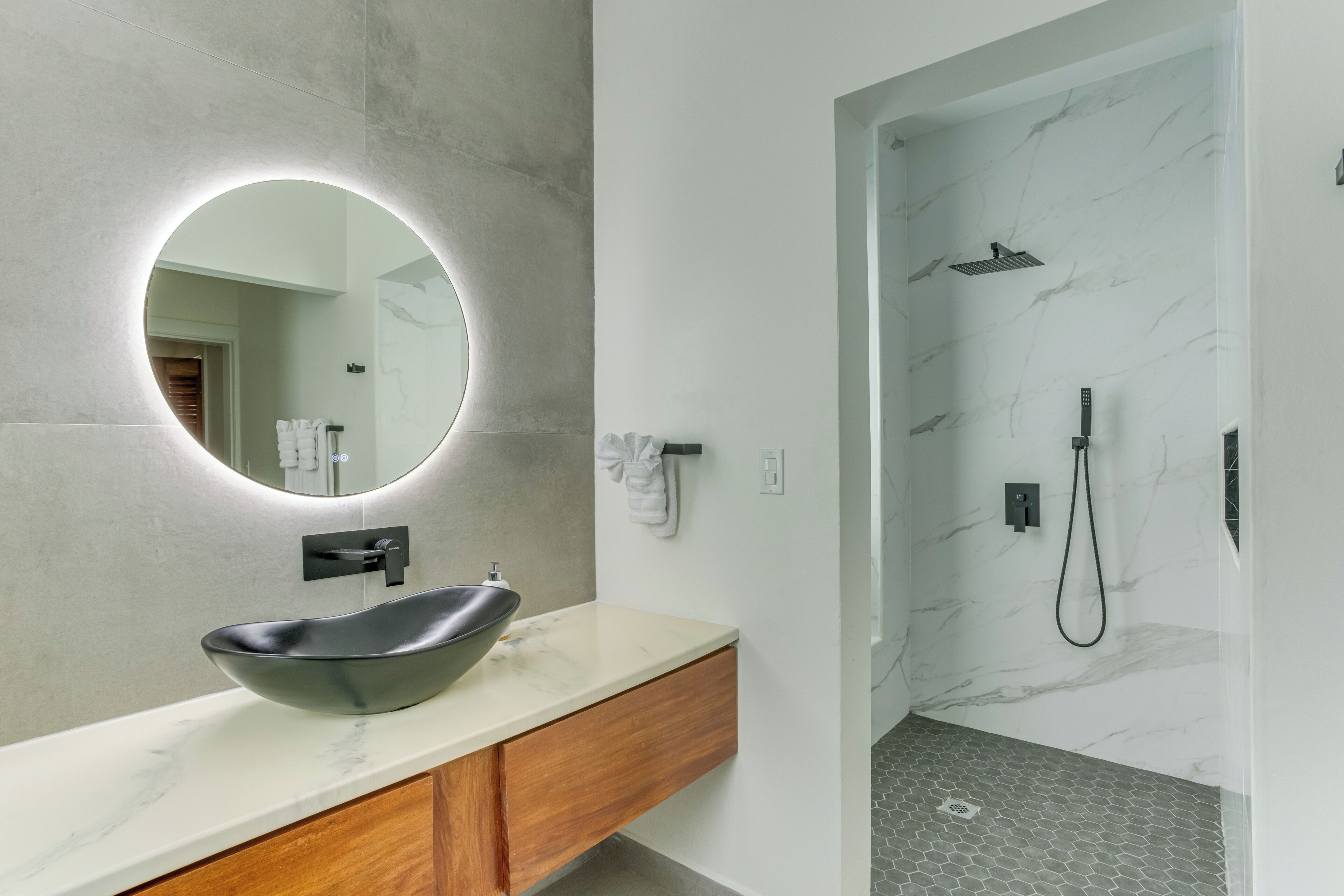 A modern bathroom with a round mirror, a black vessel sink, wooden vanity, and a shower with a marble wall.