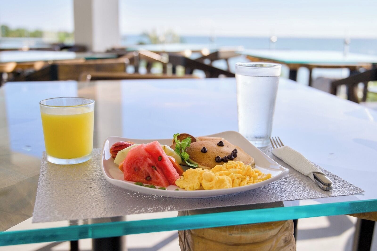 Une assiette de petit-déjeuner avec des œufs brouillés, une tranche de pastèque et un muffin, le tout posé sur une table en verre avec vue sur la mer.