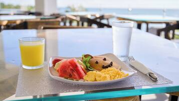 Une assiette de petit-déjeuner avec des œufs brouillés, une tranche de pastèque et un muffin, le tout posé sur une table en verre avec vue sur la mer.