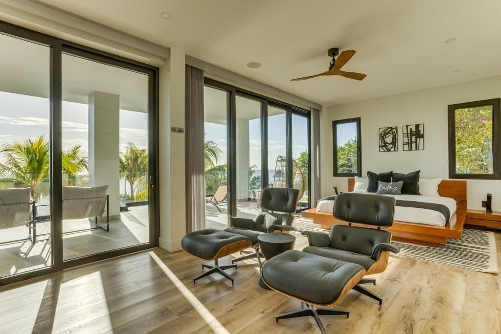 A modern bedroom with a large bed, a desk, and a chair. There is a balcony with a view of palm trees.