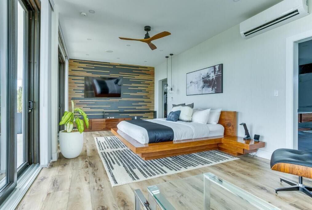 Une chambre à coucher moderne avec un lit en bois, un ventilateur de plafond, un téléviseur fixé au mur et une grande fenêtre.