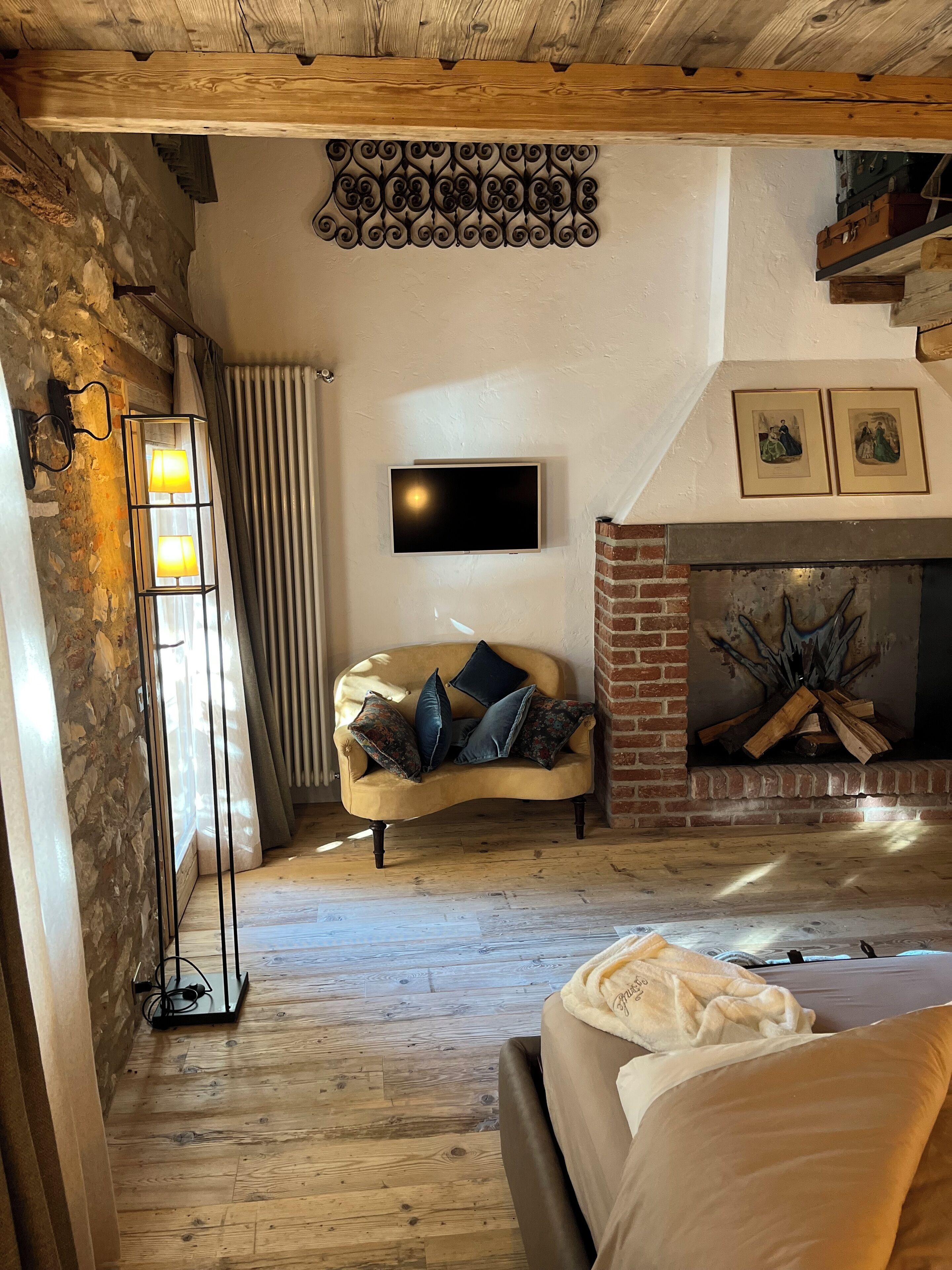 Ein gemütliches Zimmer mit Holzbalkendecke, einer Steinwand, einer Couch mit Kissen, einem Kamin und einem an der Wand befestigten Flachbildfernseher.