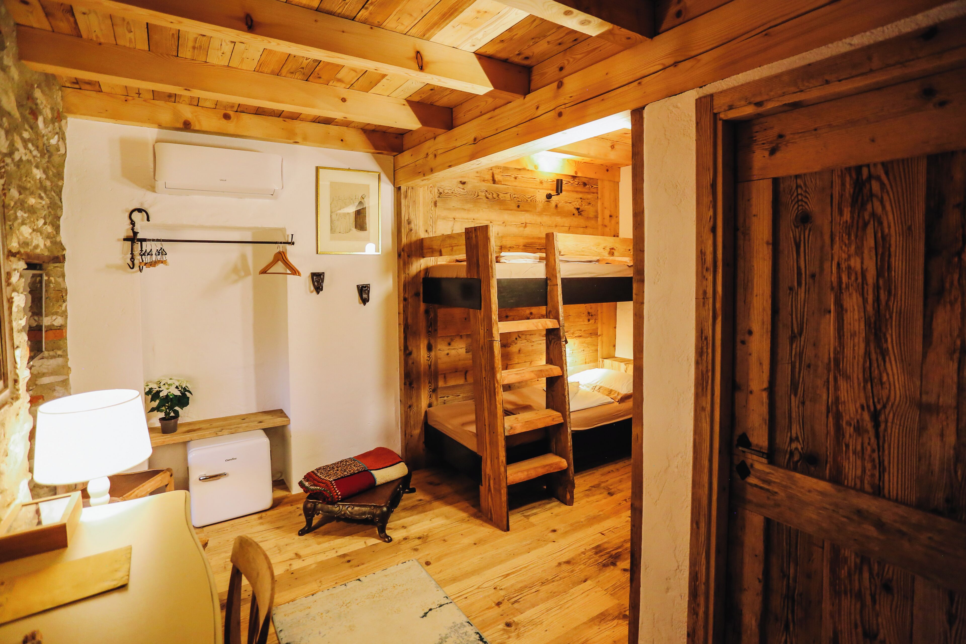 A rustic wooden room with bunk beds, a small table with a lamp, a refrigerator, and a wall-mounted air conditioner.