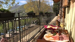 Un balcó amb una safata de fusta plena de formatge, embutits i un got de vi blanc, amb vista a un paisatge de muntanya