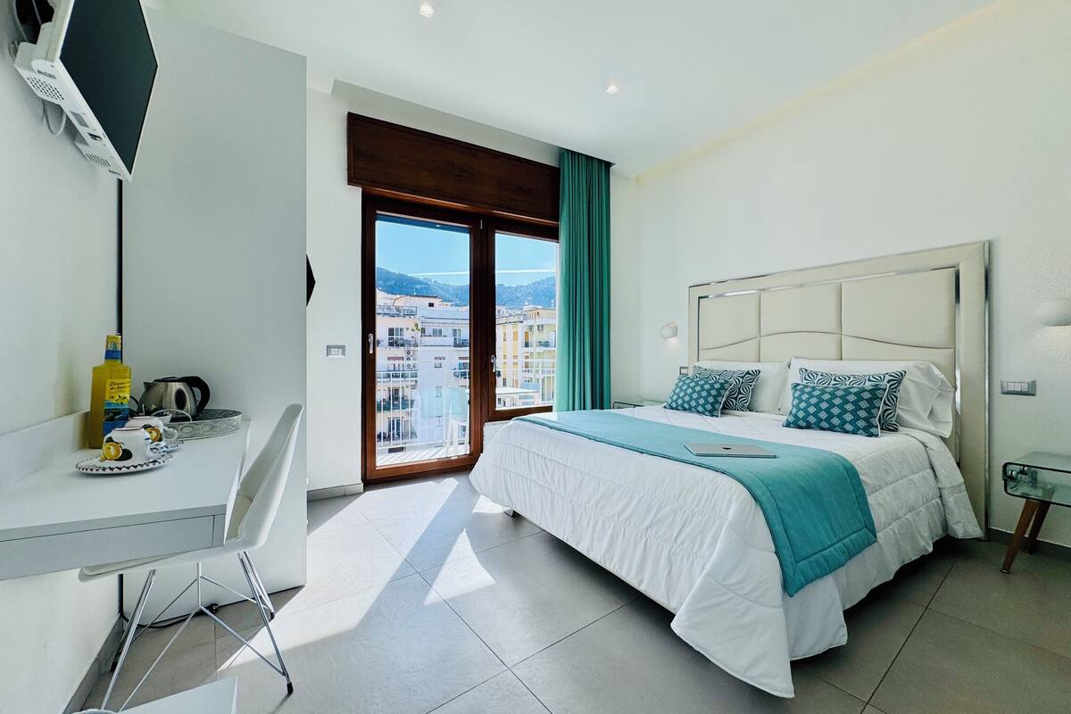 Une chambre d’hôtel moderne équipée d’un lit, d’un bureau, d’une télévision et d’un balcon offrant une vue sur la ville.