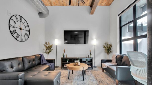 Un salon moderne avec un canapé en cuir noir, une table basse ronde en bois et un fauteuil blanc. Une grande horloge murale et un téléviseur à écran plat sont fixés au mur.