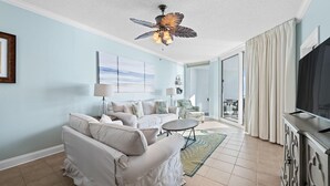 A spacious living room with a ceiling fan, a flat-screen TV, a sofa, a coffee table, and a view to the outside. - Palacio 303 (Pensacola)