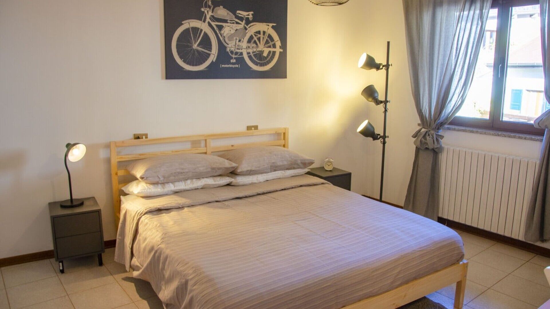 Un dormitorio con cama de madera, dos mesitas de noche, una lámpara de pared, un radiador y una ventana con cortinas.