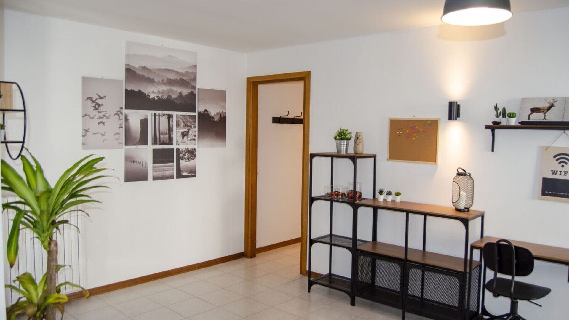 Une pièce avec une porte en bois, une étagère en métal noir ornée de plantes, un bureau avec une chaise et des tableaux accrochés au mur.