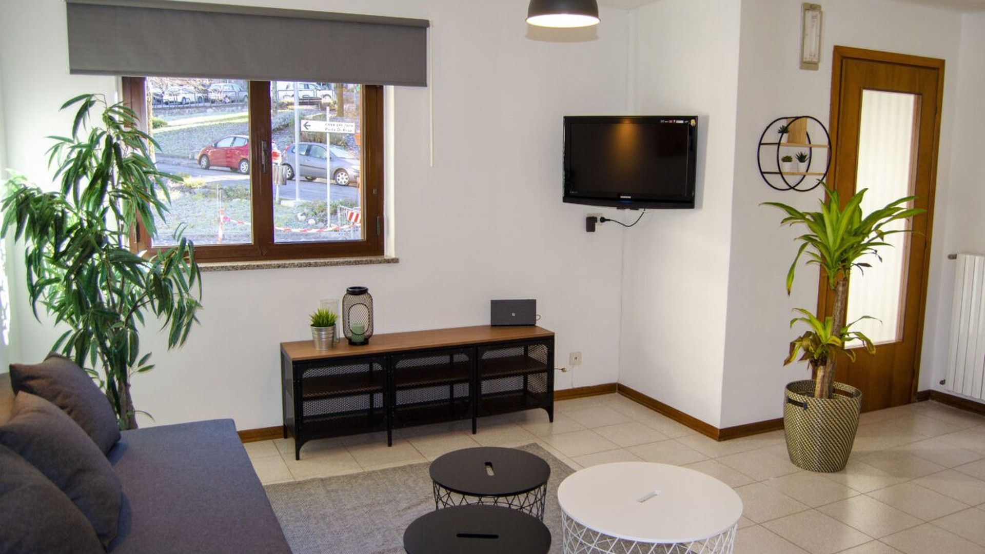 Een woonkamer met een bank, een salontafel, een aan de muur bevestigde televisie, een raam met uitzicht op geparkeerde auto's, en een kamerplant.
