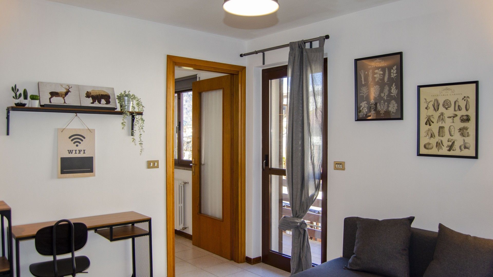 A room with a desk, a chair, a sofa, a door leading to another room, and a wall with framed botanical prints.