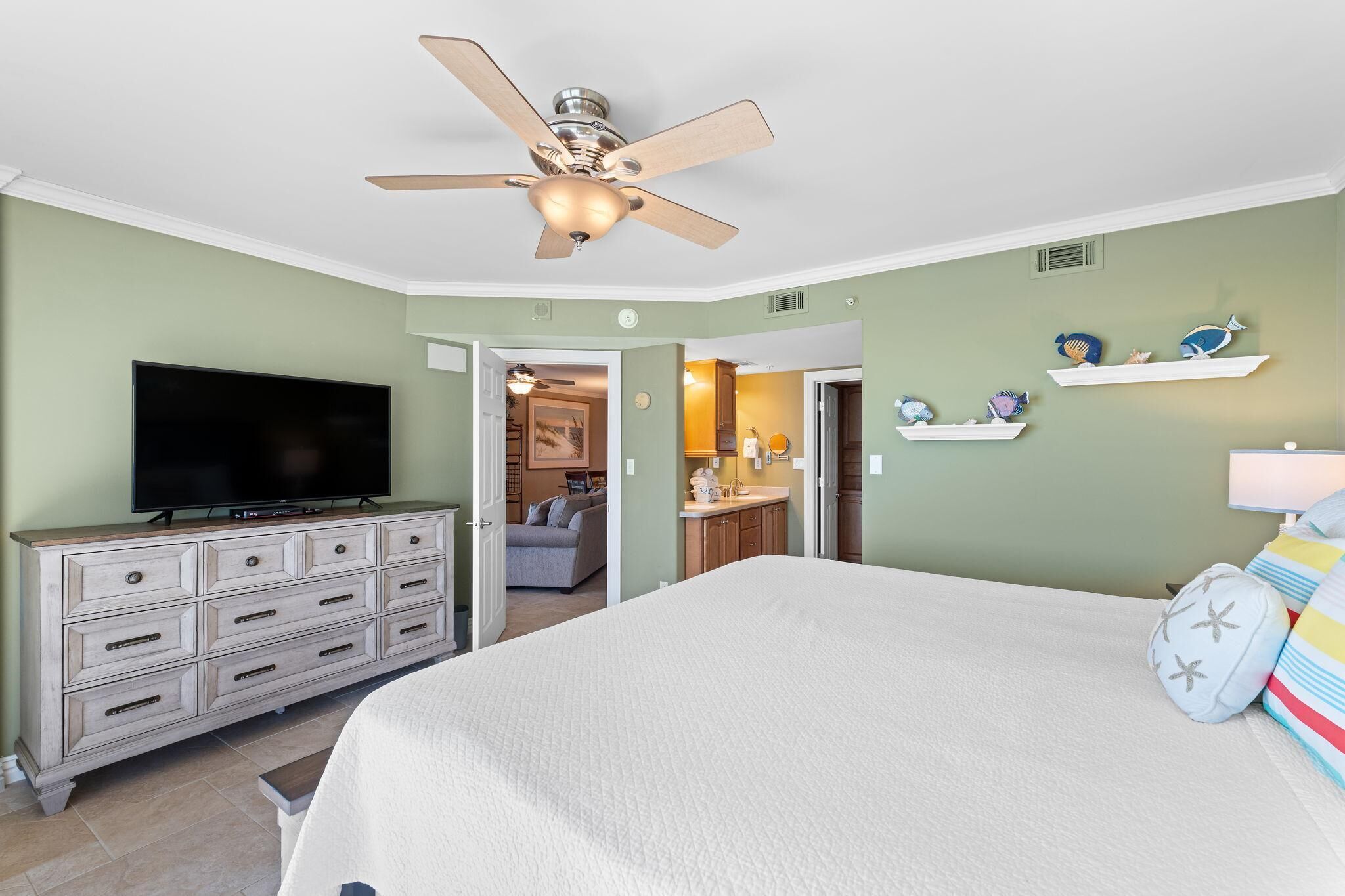 Ein Schlafzimmer mit Bett, Fernseher, Schminktisch, Deckenventilator und einem zur Toilette führenden Gang.